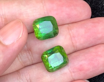 May include: Two faceted, square-cut, bright green gemstones. The gems have a vibrant, translucent quality, reflecting light. The stones are held in a hand, showcasing their color and clarity. Ideal for jewelry making or collecting.