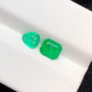Puede incluir: Dos piedras preciosas sueltas de color verde vibrante. Una piedra tiene forma de pera con una superficie facetada, mientras que la otra es una piedra rectangular de corte esmeralda con una parte superior plana y lados facetados, ambas con gran claridad.