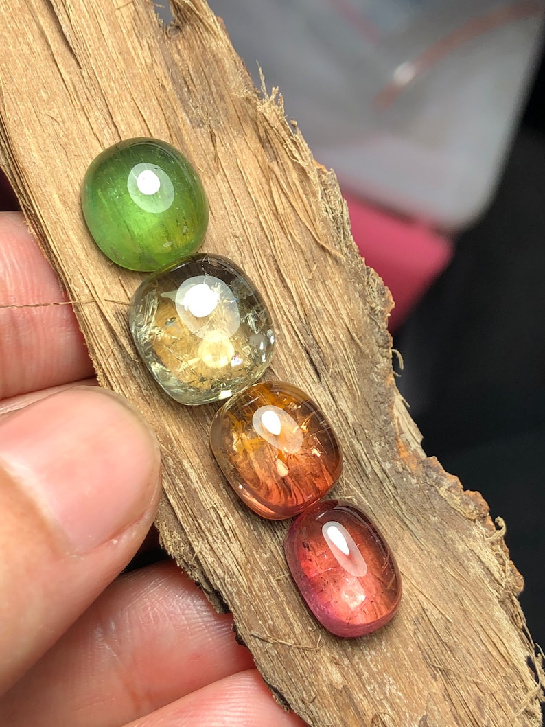 K&ouml;nnte beinhalten: F&uuml;nf ovale Edelsteine in verschiedenen Schattierungen von Gr&uuml;n, Gelb, Orange und Rosa. Die Steine sind in einer Reihe auf einem St&uuml;ck Holz angeordnet.