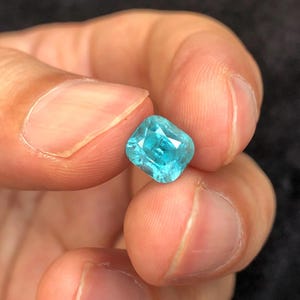 May include: A vibrant, square-cut, teal gemstone held between fingers. The gem has a faceted surface that reflects light, showcasing its clarity and color. The gemstone's corners are rounded, giving it a soft appearance.