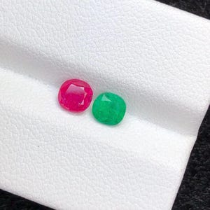 May include: A pair of gemstones, one ruby and one emerald, are displayed on a white surface. The ruby is a vibrant red, and the emerald is a rich green. Both stones have a rounded, cushion cut.