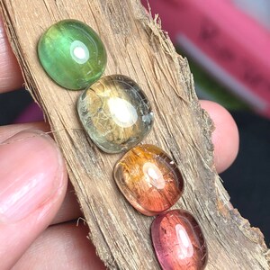 K&ouml;nnte beinhalten: F&uuml;nf Edelstein-Cabochons in verschiedenen Schattierungen von Gr&uuml;n, Gelb, Orange und Rosa. Die Steine sind in einer Reihe auf einem St&uuml;ck Holz angeordnet.