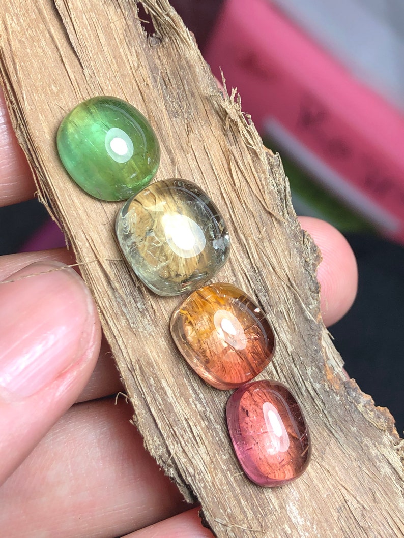 K&ouml;nnte beinhalten: F&uuml;nf Edelstein-Cabochons in verschiedenen Schattierungen von Gr&uuml;n, Gelb, Orange und Rosa. Die Steine sind in einer Reihe auf einem St&uuml;ck Holz angeordnet.