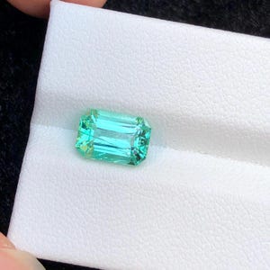 Pode incluir: Uma gema retangular com corte esmeralda de cor turquesa vibrante. A gema é exibida sobre um fundo branco, destacando sua clareza e facetas. O corte da gema permite que a luz se reflita, criando um efeito brilhante.