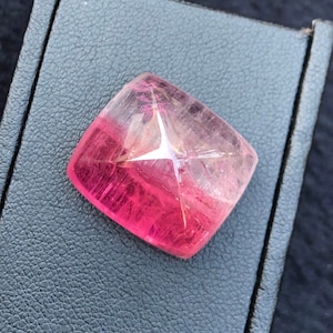 May include: A square-cut gemstone with a gradient of colors, transitioning from a deep pink at the base to a lighter pink and clear top. A star-shaped inclusion is visible within the stone. The gemstone is set on a dark gray surface.