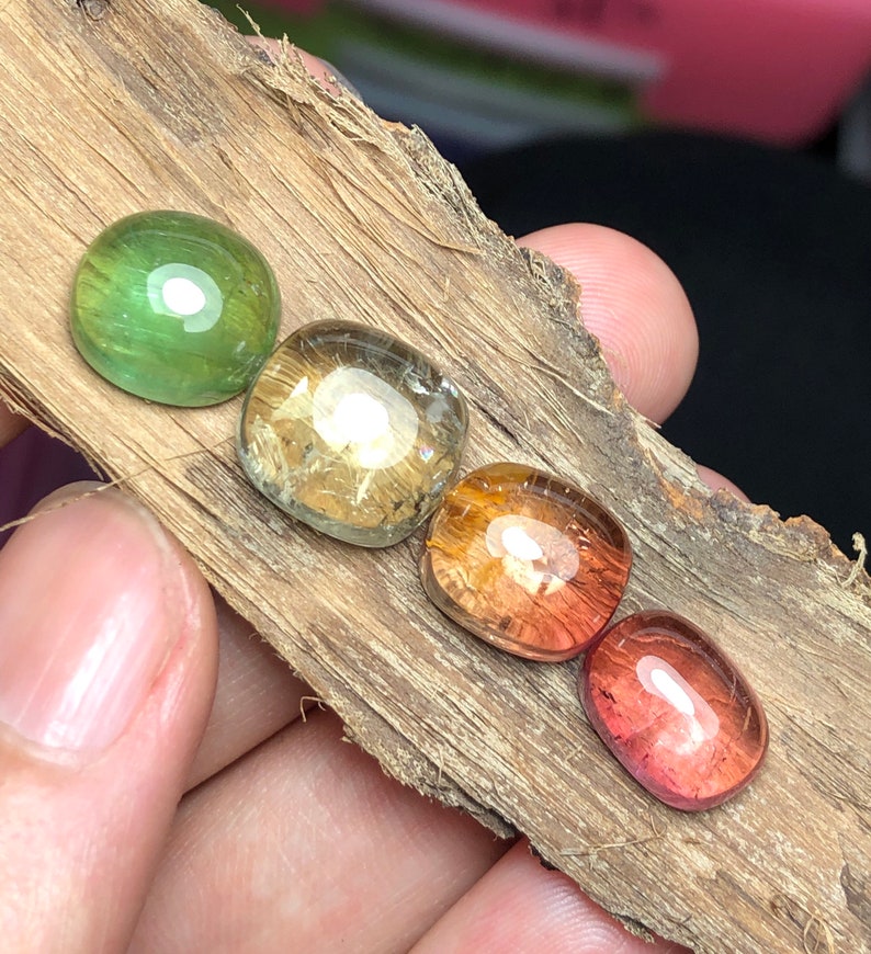 K&ouml;nnte beinhalten: Vier ovale Edelsteine in verschiedenen Farben, darunter Gr&uuml;n, Gelb, Orange und Rot, sind in einer Reihe auf einem St&uuml;ck Holz angeordnet.