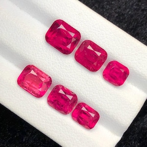 Könnte beinhalten: Sechs leuchtend rote Edelsteine im Kissenschliff. Die Edelsteine sind auf einer weißen Oberfläche angeordnet, wodurch ihre Facetten und ihre lebendige Farbe zur Geltung kommen. Die Steine variieren leicht in der Größe und haben ein poliertes, reflektierendes Aussehen.