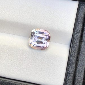 May include: A square-cut gemstone with a mix of colors, including pink, peach, and white. The gem is set against a white background within a black box, highlighting its facets and brilliance. The gemstone is a beautiful example of fine jewelry.