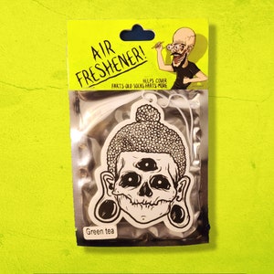 May include: A white car air freshener with a black and white illustration of a skull with three eyes and a bun on its head. The text "Air Freshener!" is printed in black on a white background. The text "Farts old socks farts more" is printed in black on a white background. The text "Helps cover" is printed in black on a white background. The text "Green tea" is printed in black on a white background.
