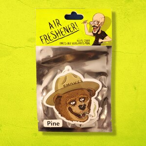 May include: A white car air freshener with a cartoon illustration of a bear wearing a ranger hat with the word "SMOKEY" on it. The bear is smoking a cigarette and has a toothy grin. The text "Pine" is printed below the image. The text "AIR FRESHENER!" is printed at the top of the package. The text "FARTS-OLO SOCKS FARTS MORE" is printed below the image. The text "HELPS COVER" is printed above the image.
