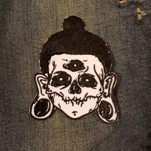 May include: Black and white embroidered patch of a skull with three eyes and large ear gauges. The skull is wearing a black bun on its head.