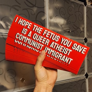 Pode incluir: Uma pilha de adesivos vermelhos para pára-choques com texto branco que diz "Espero que o feto que você salva seja um ateu queer comunista imigrante".
