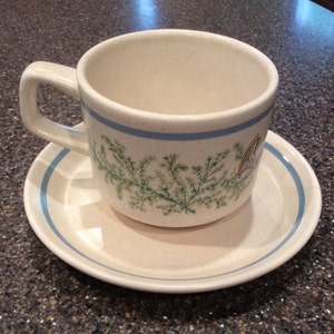 May include: A white ceramic teacup and saucer with a blue trim and a floral design featuring green leaves and a butterfly.