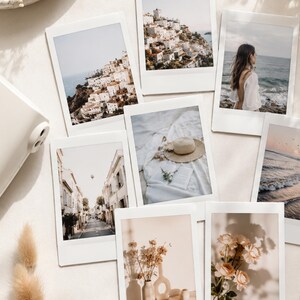 May include: A white instant camera positioned next to a collection of seven rectangular instant film prints featuring various scenes including coastal towns, a beach, a straw hat on fabric, a street view, dried flowers, and light-coloured roses.