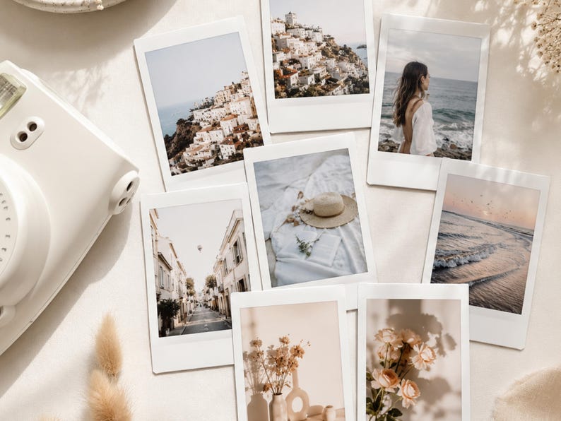 May include: A white instant camera positioned next to a collection of seven rectangular instant film prints featuring various scenes including coastal towns, a beach, a straw hat on fabric, a street view, dried flowers, and light-coloured roses.