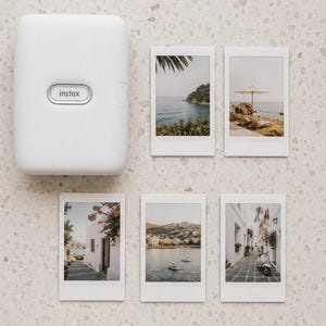 May include: A white Instax portable photo printer with a textured surface and five rectangular instant film prints featuring various travel scenes, including coastal landscapes, a beach umbrella, white buildings, and a scooter on a stone path.