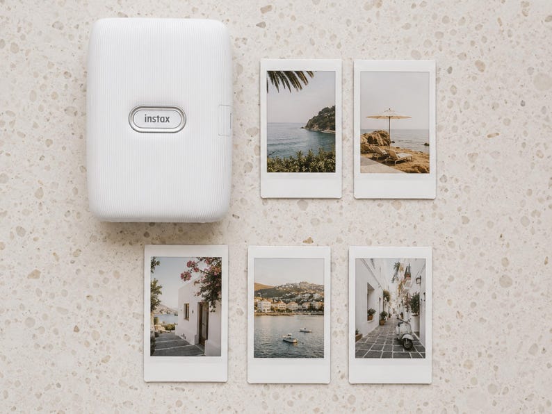 May include: A white Instax portable photo printer with a textured surface and five rectangular instant film prints featuring various travel scenes, including coastal landscapes, a beach umbrella, white buildings, and a scooter on a stone path.