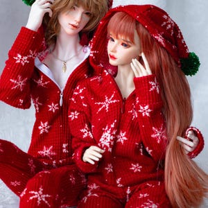 May include: Two dolls in matching red onesie pyjamas with white snowflake patterns. The pyjamas have hoods with green pom-poms. The dolls have long hair and are posed in a studio setting.