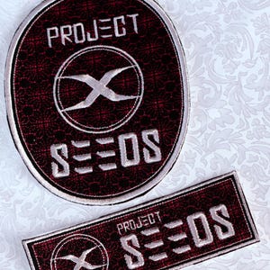 Puede incluir: Dos parches bordados con el texto "PROJECT SEEDS". Uno es ovalado, el otro rectangular. Ambos tienen un fondo burdeos con un diseño y borde bordados en blanco. El diseño incluye una "X" estilizada dentro de un círculo.