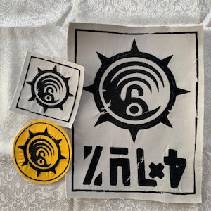Puede incluir: Tres parches de tela con un emblema circular negro con puntas radiantes y líneas curvas. Un parche rectangular blanco grande incluye el emblema negro sobre texto negro estilizado. Un parche cuadrado blanco pequeño y uno circular amarillo también presentan el emblema.