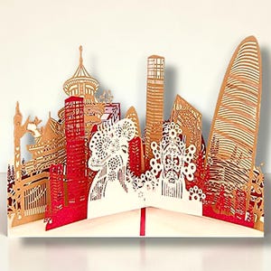May include: A three-dimensional paper art piece depicting a cityscape. The design features buildings in shades of red, tan, and white, with intricate cut-out details. The card is folded and stands upright, showcasing the architectural elements.