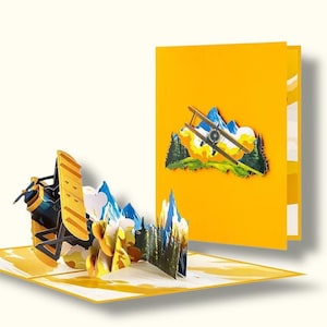 May include: A yellow pop-up card with a biplane and mountain scene. The open card displays a 3D airplane design with blue mountains and yellow accents. The closed card shows a similar design. The card is made of paper.