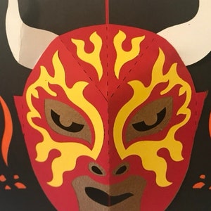 May include: A red and yellow luchador mask with white horns and a brown mouth. The mask has a flame design and is set against a black background with orange flame accents. The mask is a paper craft.