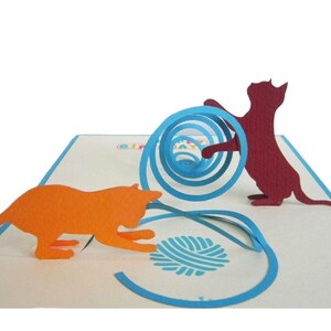 Op de afbeelding: Een pop-up wenskaart met twee katten silhouetten. Een oranje kat reikt naar een blauwe bol wol, terwijl een bordeauxrode kat interactie heeft met een blauwe spiraal. De kaart staat op een witte achtergrond met blauwe accenten.