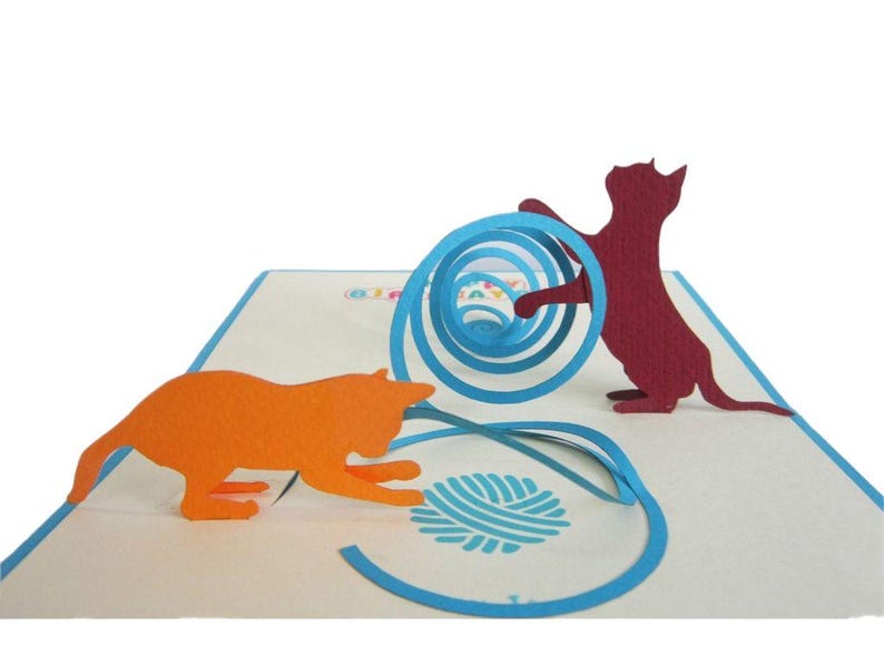 Op de afbeelding: Een pop-up wenskaart met twee katten silhouetten. Een oranje kat reikt naar een blauwe bol wol, terwijl een bordeauxrode kat interactie heeft met een blauwe spiraal. De kaart staat op een witte achtergrond met blauwe accenten.