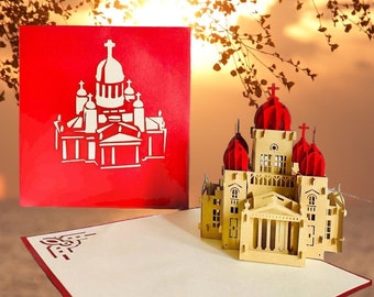 Puede incluir: Una tarjeta de papel pop-up roja y beige con un modelo 3D detallado de una iglesia con techos rojos y una fachada blanca. La tarjeta está cerrada y muestra una silueta de la iglesia en la parte delantera.