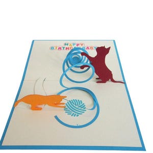 Op de afbeelding: Een pop-up verjaardagskaart met twee katten silhouetten die met een bolletje wol spelen. De kaart heeft een blauwe rand en de woorden "HAPPY BIRTHDAY BABY" in kleurrijke letters.