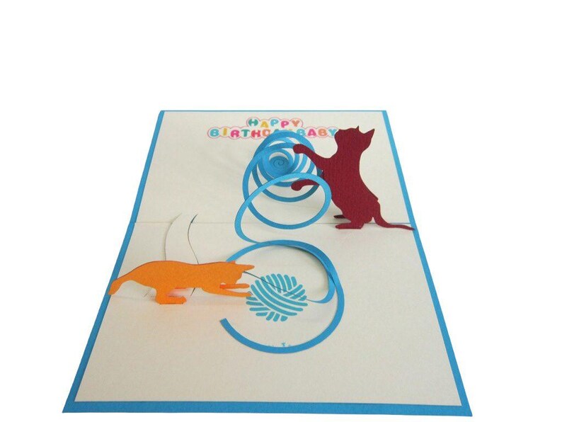 Op de afbeelding: Een pop-up verjaardagskaart met twee katten silhouetten die met een bolletje wol spelen. De kaart heeft een blauwe rand en de woorden "HAPPY BIRTHDAY BABY" in kleurrijke letters.