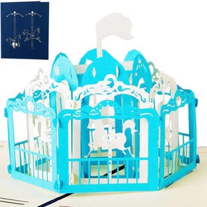 May include: A pop-up greeting card featuring a carousel design. The card is primarily light blue and white, with intricate cut-out details of horses and decorative elements. A smaller card on the left shows a carousel design in gold on a dark blue background.