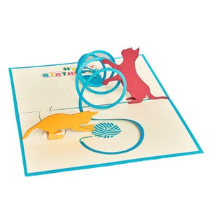 Op de afbeelding: Pop-up verjaardagskaart met twee katten silhouetten die met een bolletje wol spelen. E&eacute;n kat is oranje, de andere rood. De kaart heeft een blauwe rand en de woorden "HAPPY BIRTHAY" zijn zichtbaar.