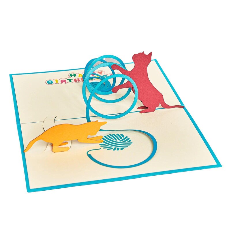 Op de afbeelding: Pop-up verjaardagskaart met twee katten silhouetten die met een bolletje wol spelen. E&eacute;n kat is oranje, de andere rood. De kaart heeft een blauwe rand en de woorden "HAPPY BIRTHAY" zijn zichtbaar.