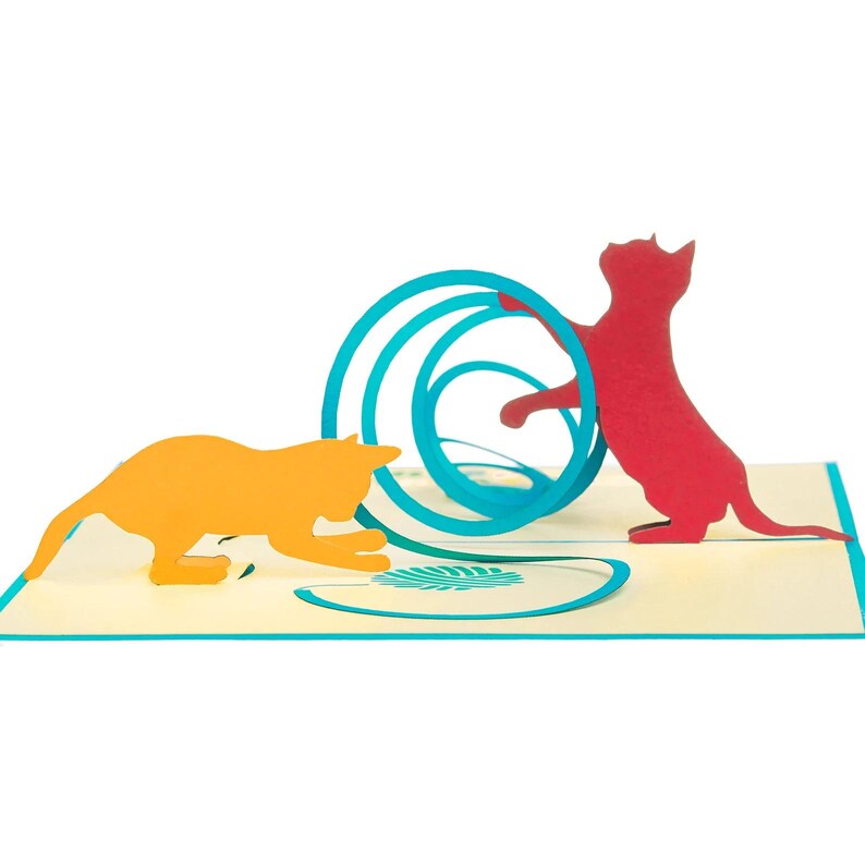 Op de afbeelding: Een pop-up wenskaart met twee katten silhouetten. Een oranje kat staat links en een rode kat rechts, die interageren met een blauw cirkelvormig ontwerp. De kaart staat op een lichtgele achtergrond.