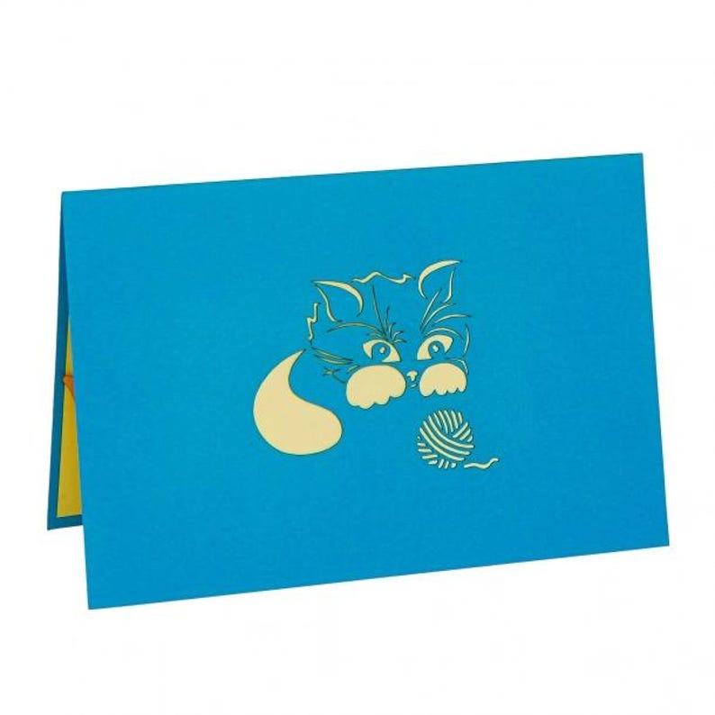 Op de afbeelding: Blauwe wenskaart met een pop-up ontwerp met een kat die met een bolletje wol speelt. De kat en het garen zijn lichtgeel en contrasteren met de blauwe achtergrond. De kaart is open en toont een gele binnenkant.