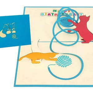 Op de afbeelding: Een pop-up verjaardagskaart met een katten thema. De kaart heeft een blauwe rand en een blauwe omslag met een kat en garen ontwerp. Binnenin spelen een rode en oranje kat met blauw garen, met de woorden "HAPPY BIRTHDAY" erboven.