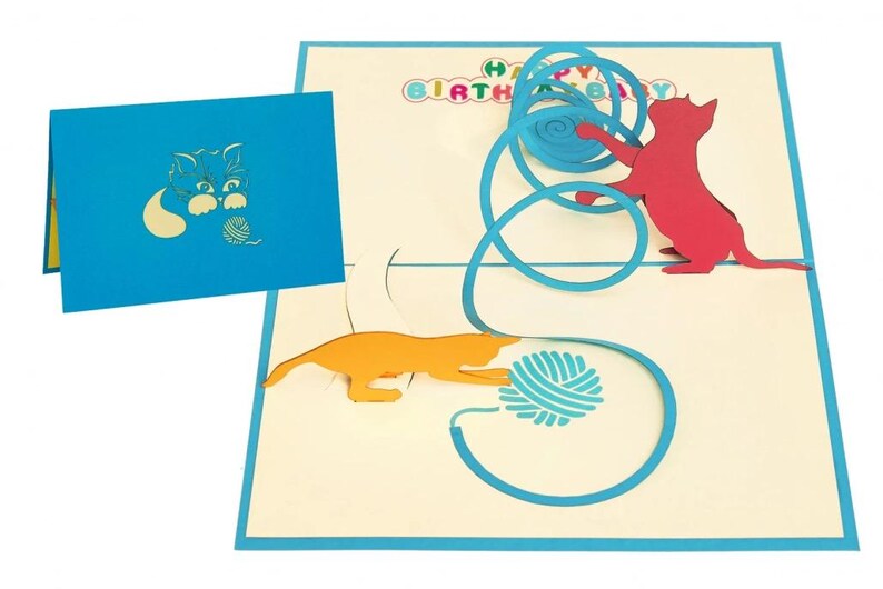 Op de afbeelding: Een pop-up verjaardagskaart met een katten thema. De kaart heeft een blauwe rand en een blauwe omslag met een kat en garen ontwerp. Binnenin spelen een rode en oranje kat met blauw garen, met de woorden "HAPPY BIRTHDAY" erboven.