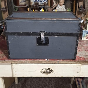 May include: A vintage gray trunk with black metal trim and a latch. The trunk is rectangular and sits on a white table with a drawer. The background includes a license plate and other antique items.