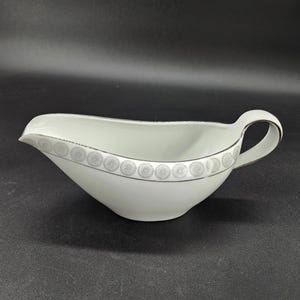 May include: White gravy boat with a handle and a spout. The rim is decorated with a repeating silver floral pattern. The gravy boat is made of porcelain and is on a black surface.