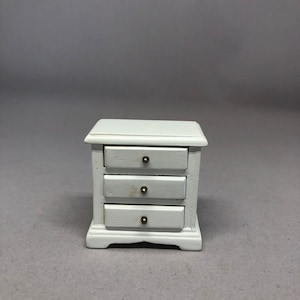 May include: White wooden dresser with three drawers and brass knobs. The dresser is a miniature piece of furniture.