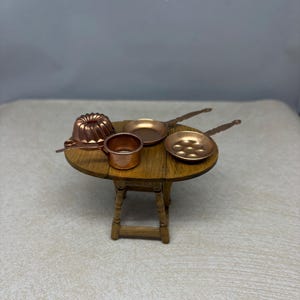 May include: A miniature wooden drop-leaf table holding a collection of small copper-colored cookware, including a fluted bundt mold, a deep saucepan, a flat skillet, and a small pan with multiple circular indentations on the surface.