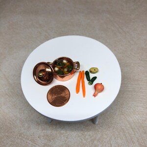 May include: A miniature copper pot with a lid filled with green vegetables and white objects, along with two orange carrots, a green cucumber, a small yellow onion, and a brown penny on a white round table with three legs.
