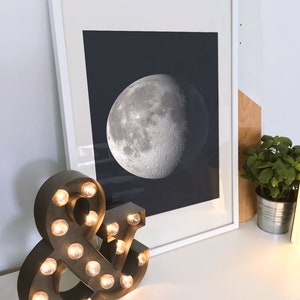May include: A framed print of a black and white image of the moon, hanging on a white wall above a brown and black light-up ampersand sign.