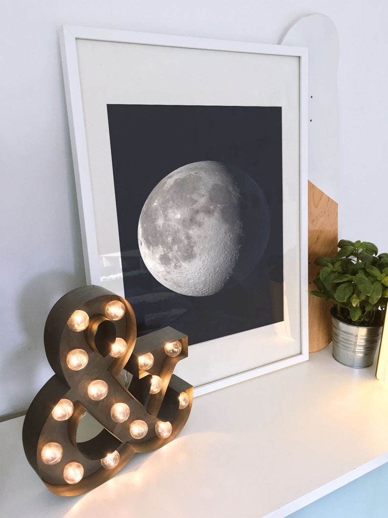 May include: A framed print of a black and white image of the moon, hanging on a white wall above a brown and black light-up ampersand sign.