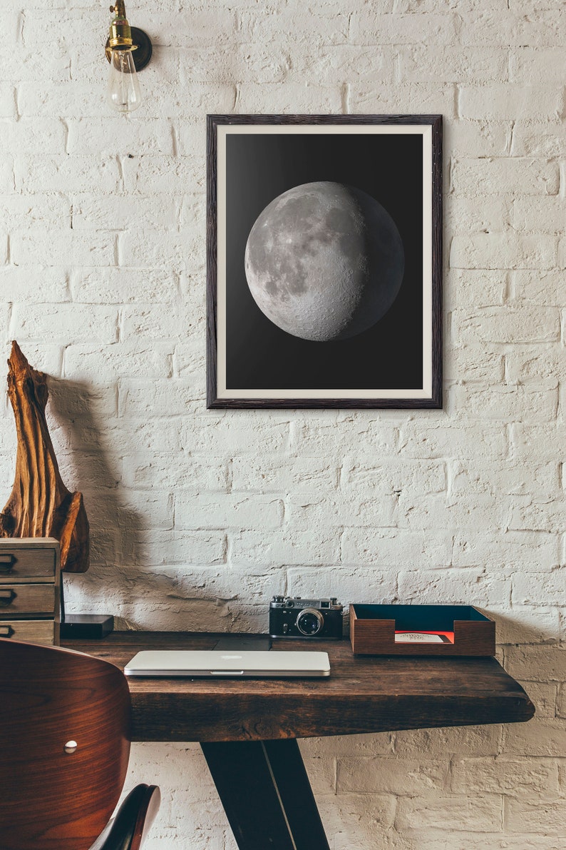 May include: A framed black and white photograph of the moon, hanging on a white brick wall above a wooden desk with a laptop, camera, and a wooden tray.