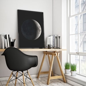 May include: A black and white photo of a crescent moon hanging on a wall above a wooden desk with a black chair and a wooden easel.