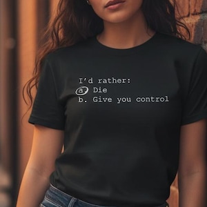 May include: Black crew neck t-shirt with white text that reads "I'd rather: Die b. Give you control". The shirt is paired with blue jeans. The model is leaning against a brick wall.