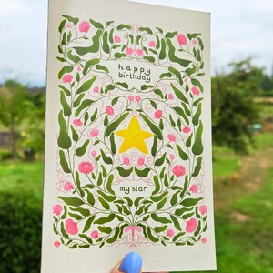 Pu&ograve; includere: Biglietto di auguri con un design floreale e le parole "buon compleanno" e "my star". Il biglietto presenta una stella gialla al centro, circondata da fiori rosa e foglie verdi. Lo sfondo &egrave; color crema chiaro.